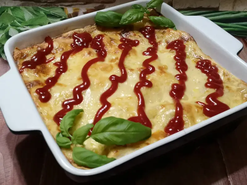 Pyszne, zapieczone fit lasagne w białym naczyniu, polane ketchupem i ozdobione listkami bazylii.